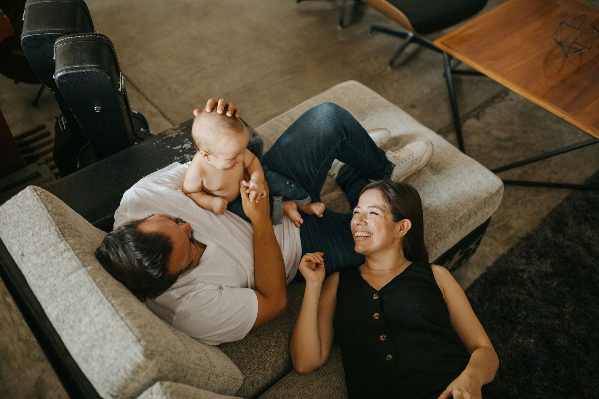 Family of three on a couch: adult man, adult woman, and baby. The baby sits on the man's lap. All are smiling.