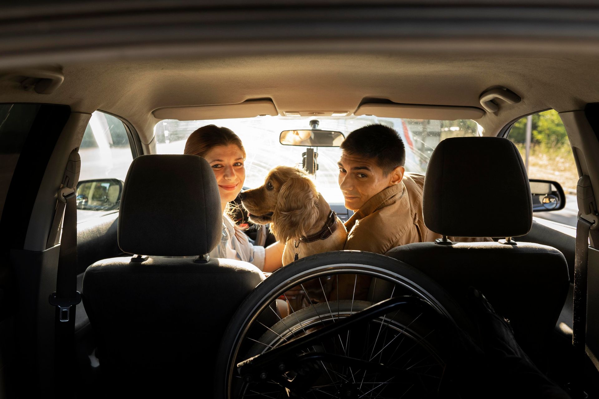 Two people and dog in a car; wheelchair in trunk. Smiling, sunny day.