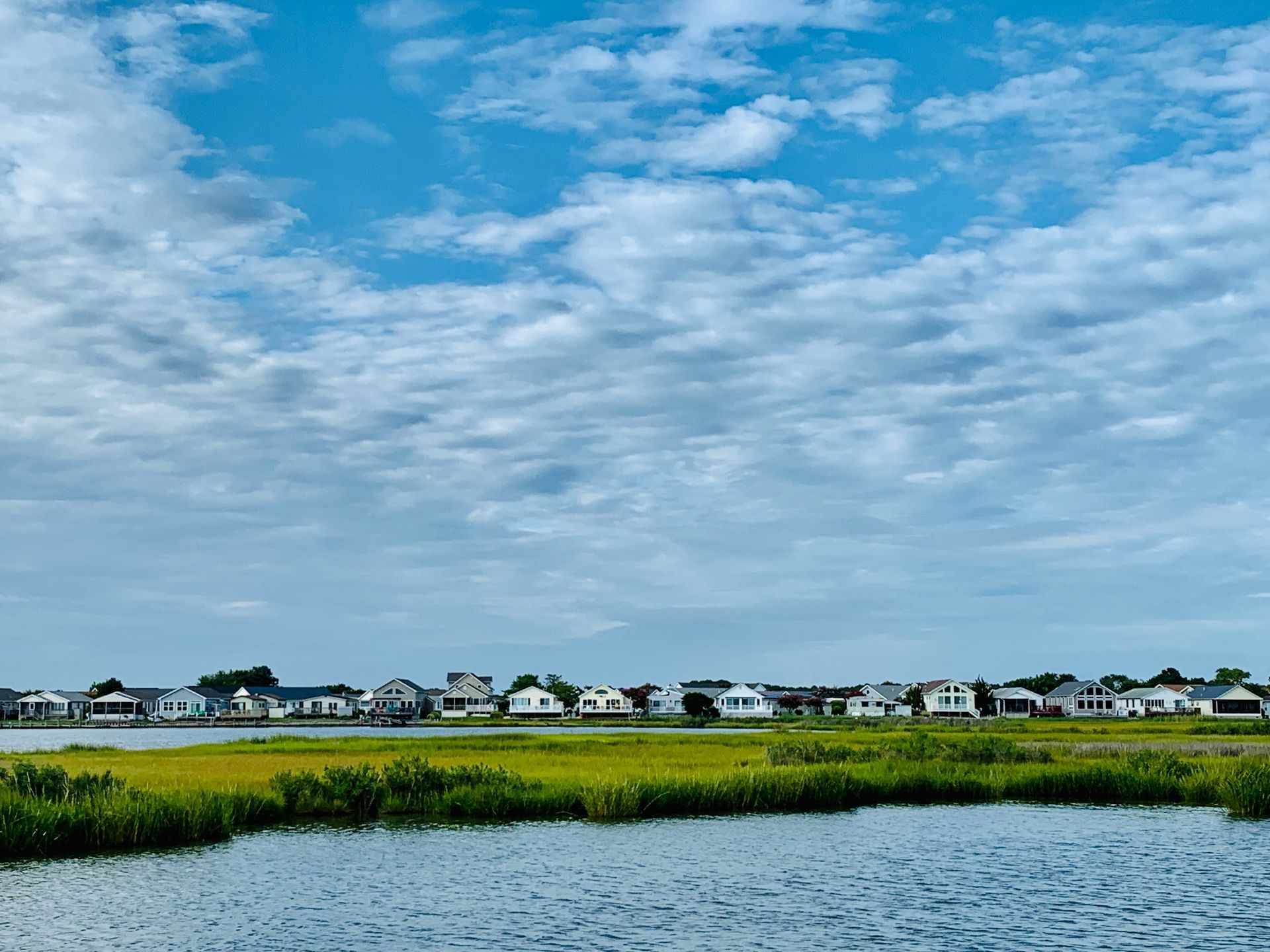 Waterfront houses along a grassy marsh under a partly cloudy blue sky.