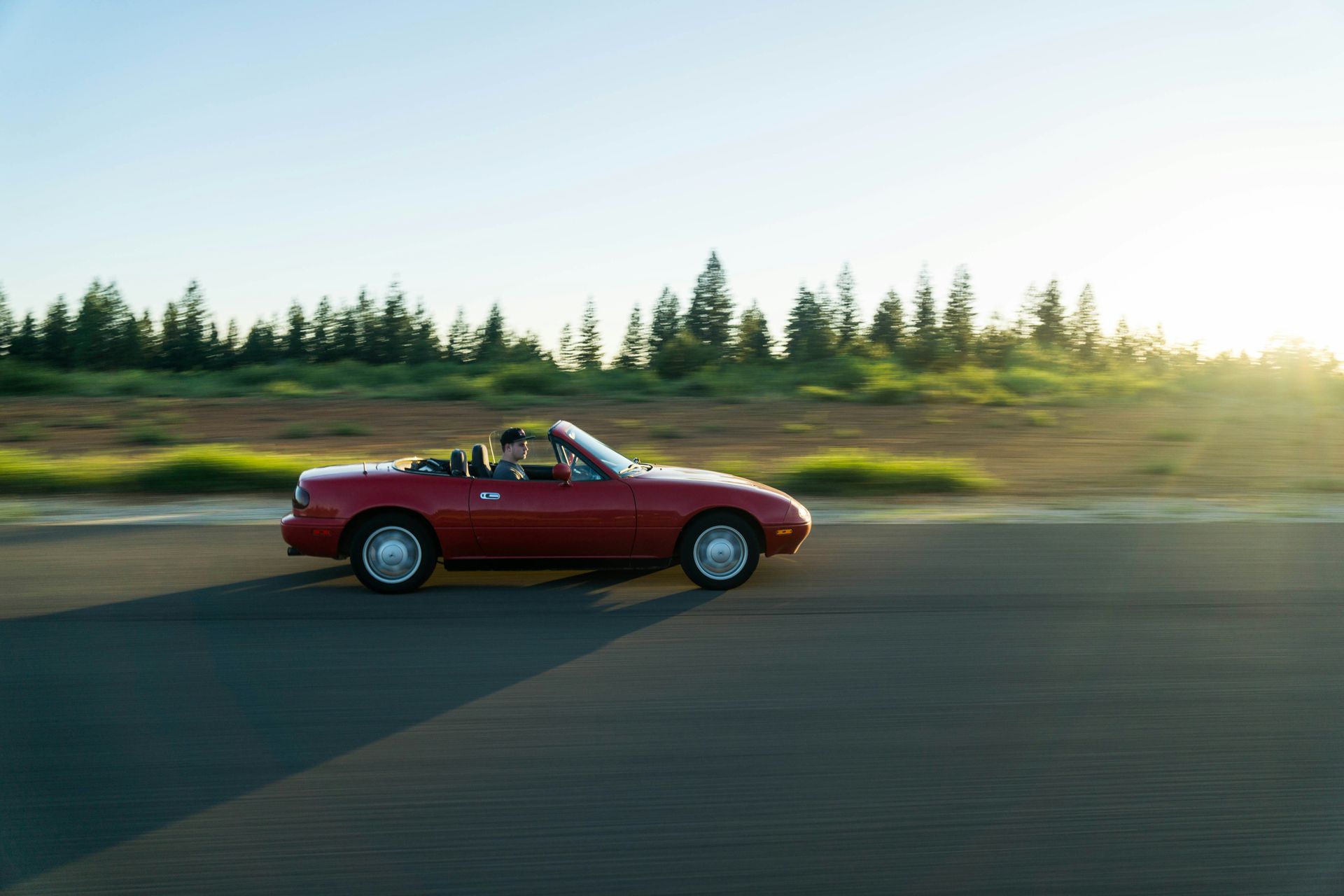 Red convertible car driving on a road, with trees in the background, sun setting.