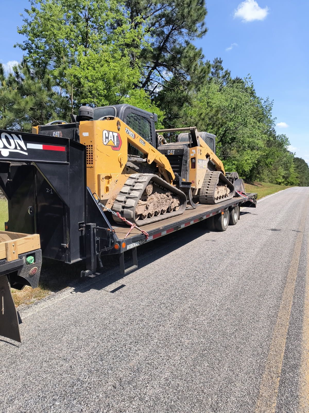 Two yellow CAT skid steer loaders loaded onto a black flatbed trailer parked on the side of a rural road.