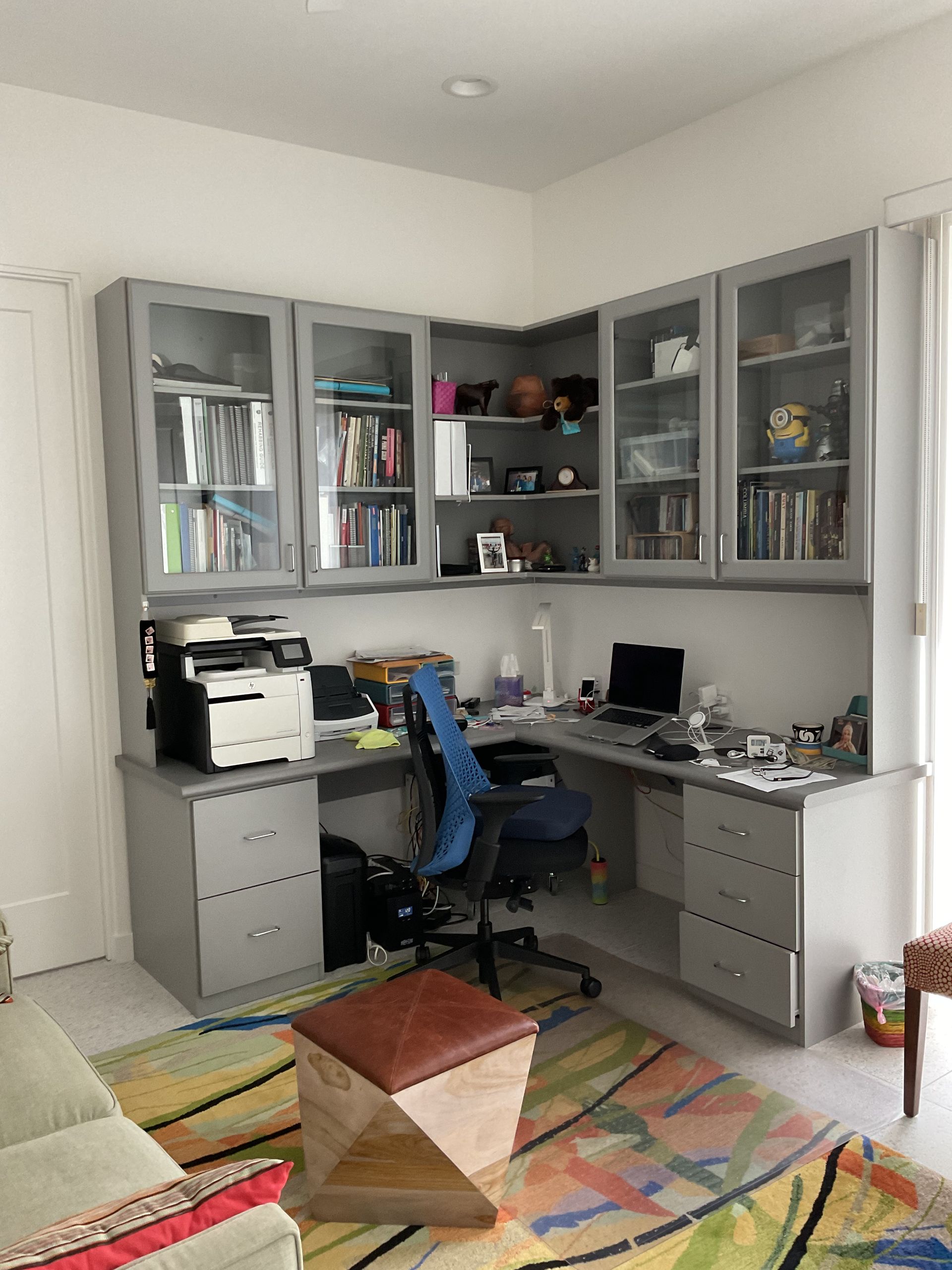 L-shaped home office with gray cabinets, desk, and chair. Books, printer, and laptop present. Colorful rug.