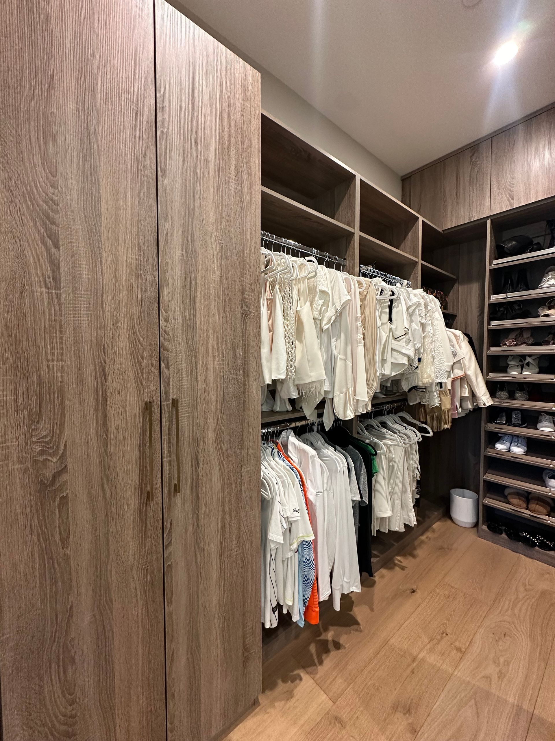 A walk-in closet with wooden shelves and hanging clothes. Light wooden floors and walls.