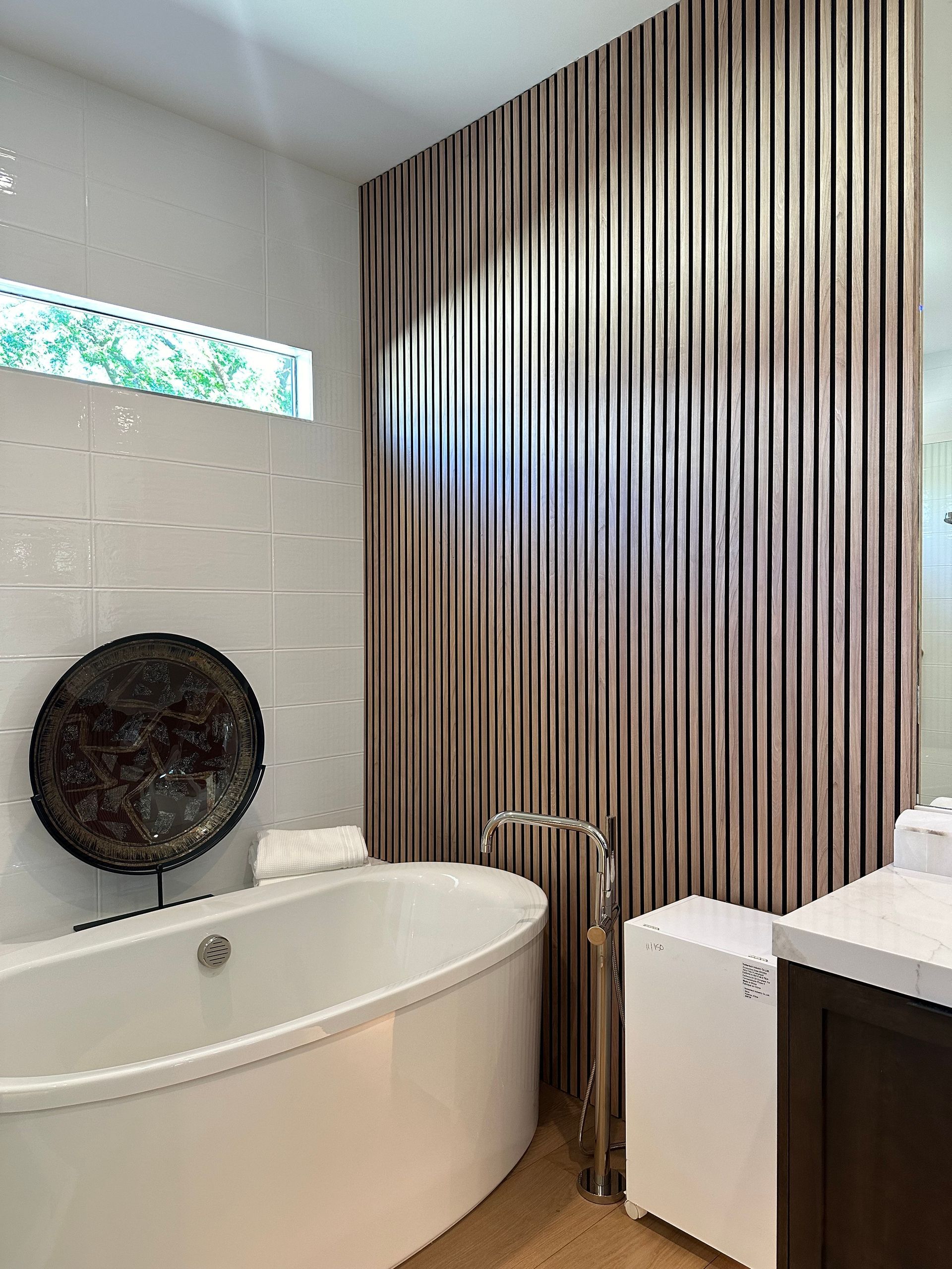 Oval bathtub in a modern bathroom with textured wall, small window, and brown vertical panels.