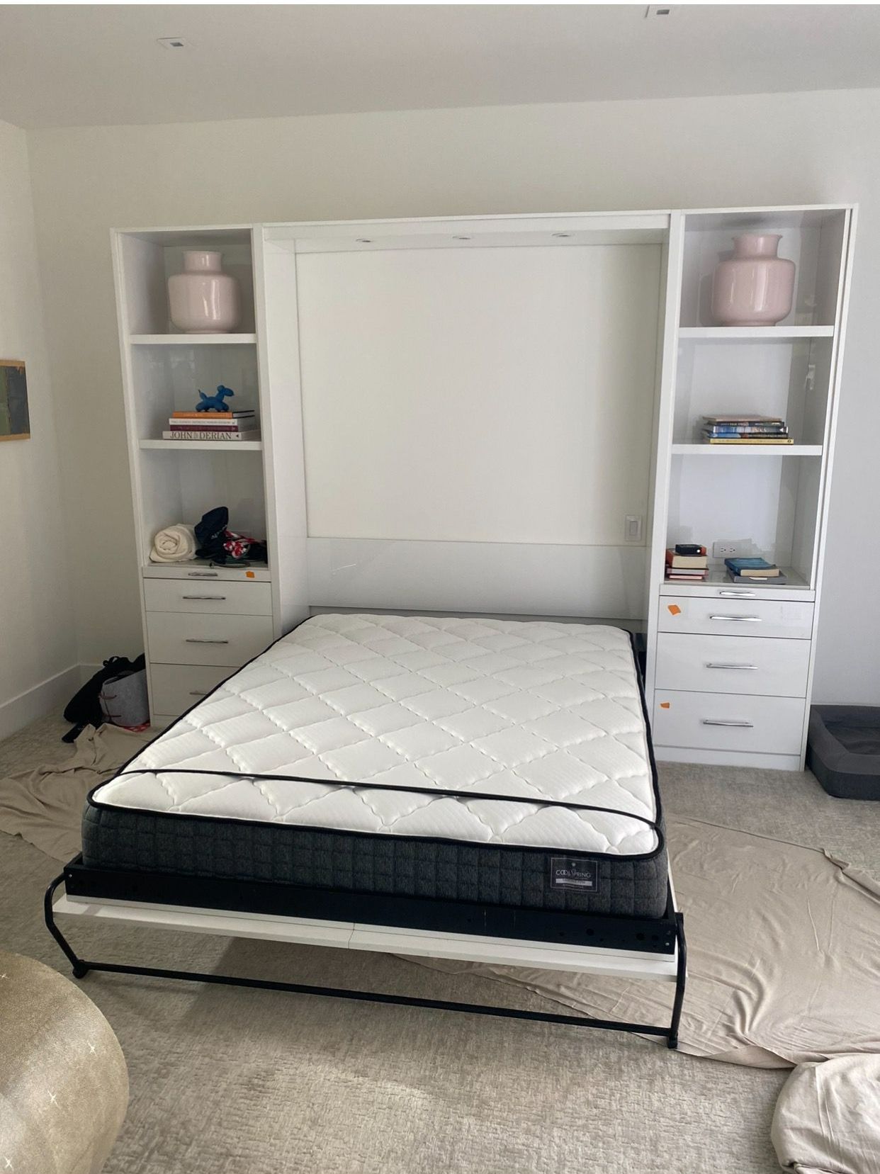 White wall bed with mattress, flanked by shelves with pink vases and drawers.
