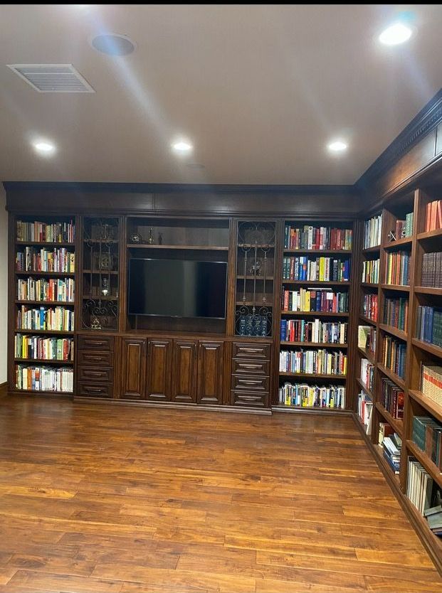 A library room with dark wood bookshelves filled with books, surrounding a TV. Wooden floor.