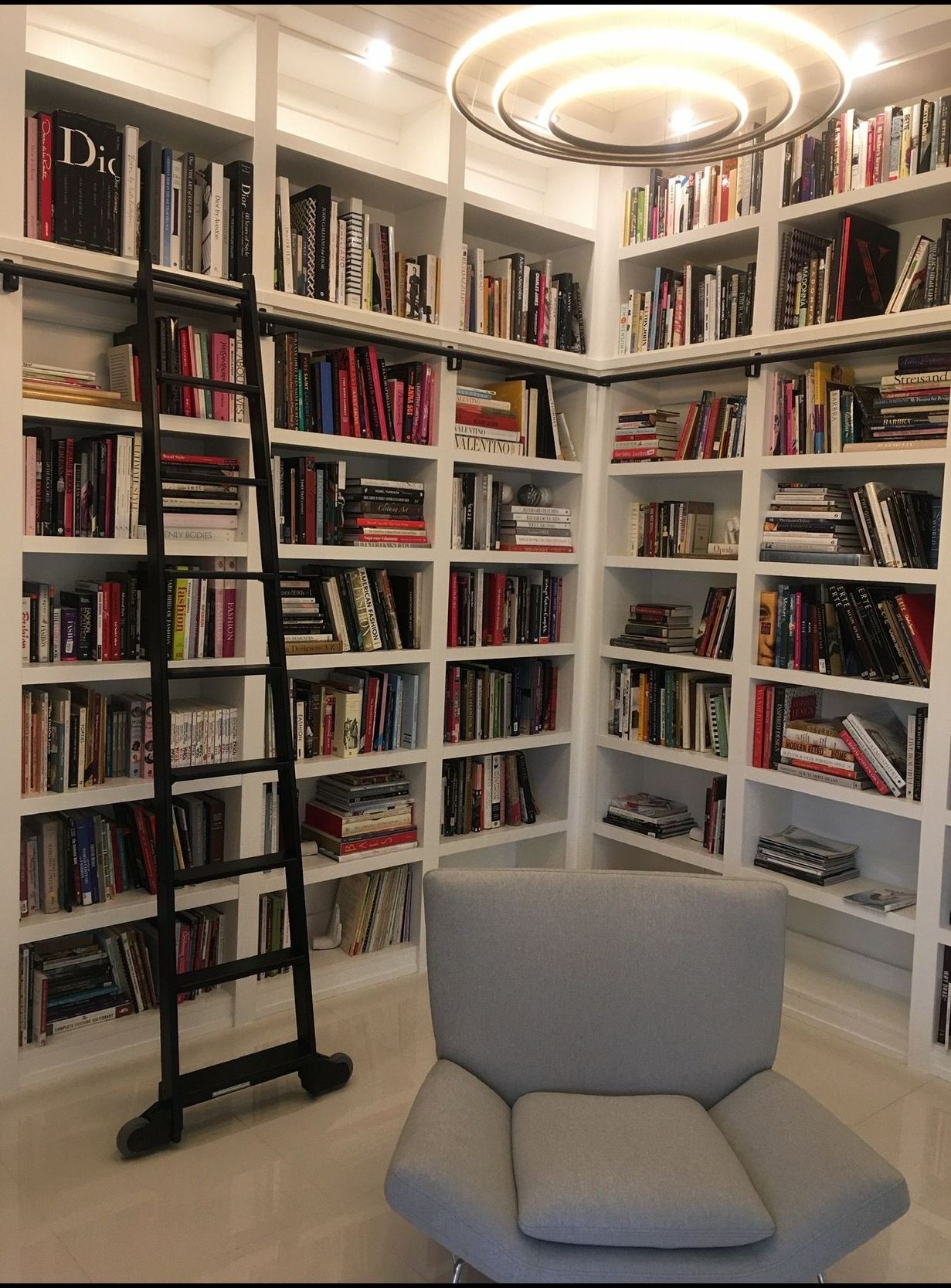A cozy library with white bookshelves, a black ladder, gray armchair, and a circular light fixture.