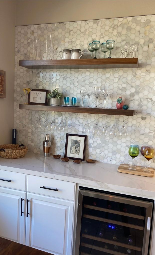 Built-in home bar with white cabinets, marble hexagon tile backsplash, and wooden shelves displaying glassware and bottles.