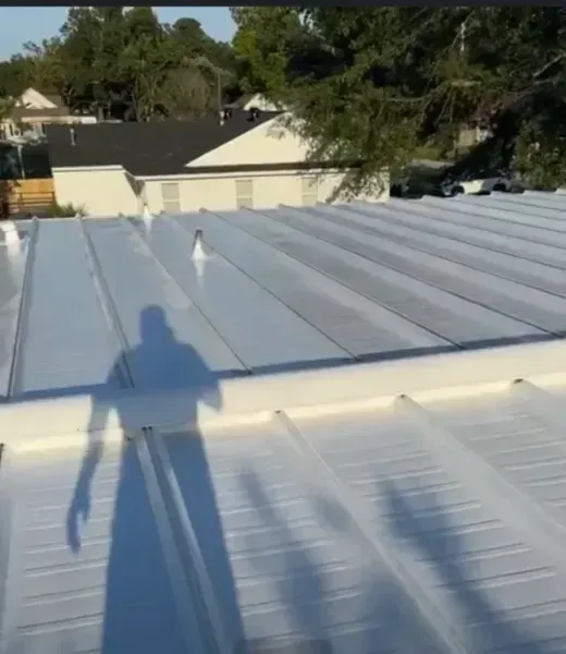 A high-angle view of a newly coated white metal roof with standing seams, seen under bright sunlight with a human shadow.