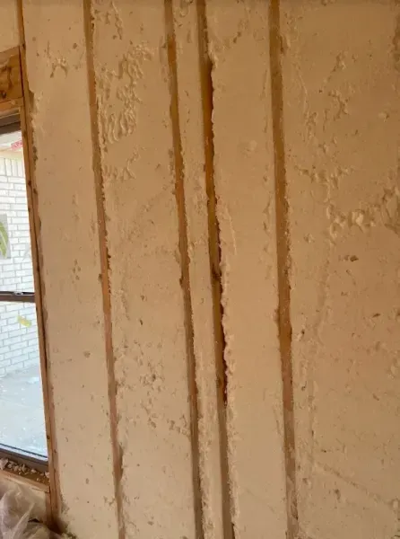 An interior wall featuring exposed vertical wooden studs covered with textured, light-colored plaster or foam insulation.