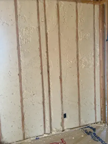 Wall studs filled with beige spray foam insulation, with an electrical outlet visible in the center.