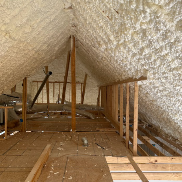 An attic interior with wooden beams and floorboards, featuring walls and ceiling coated in thick, white spray foam.
