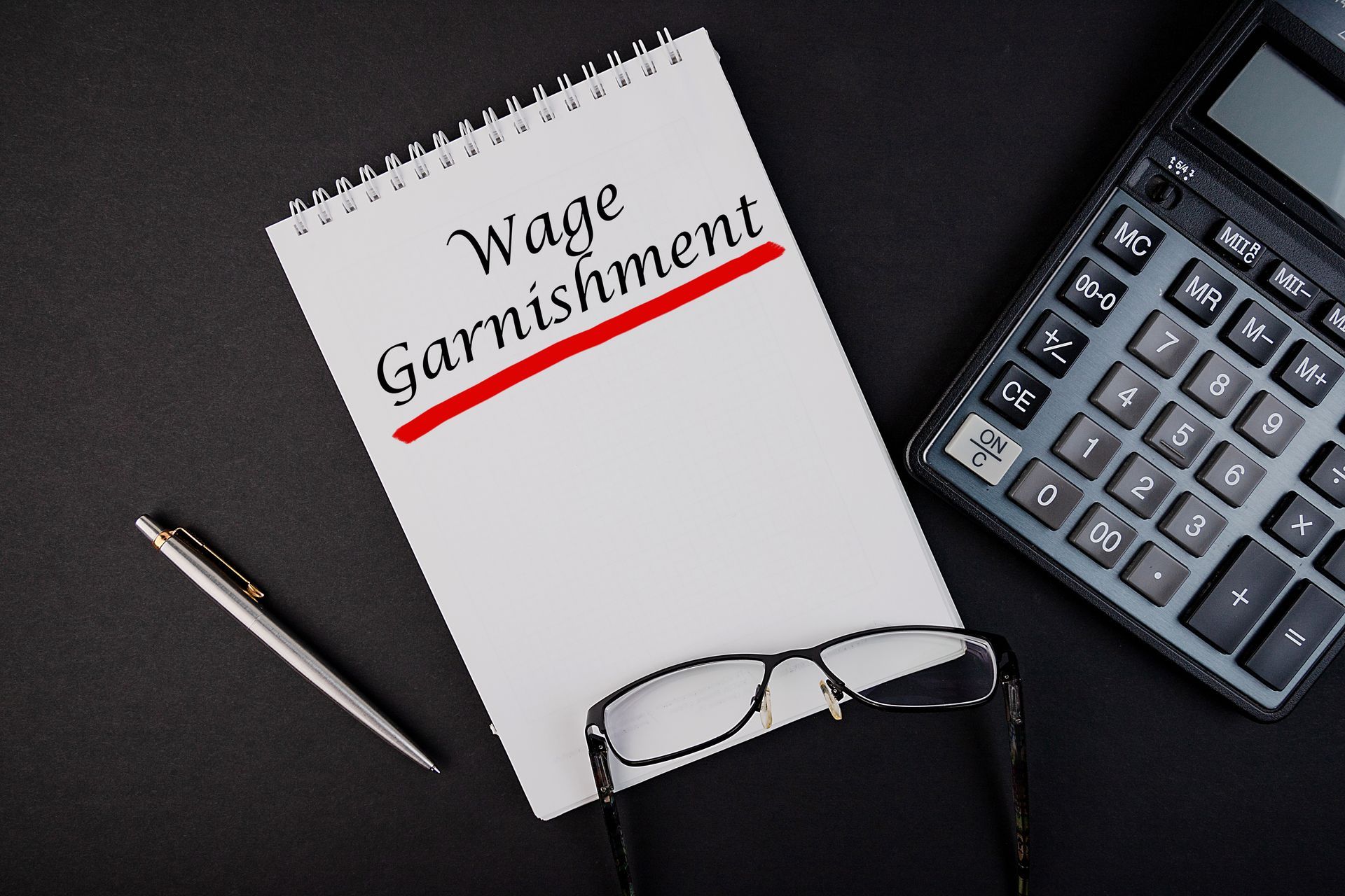 Wage Garnishment Written in a Notebook Beside a Pen and Calculator. Wage Garnishment Written in a Notebook Beside a Pen and Calculator.