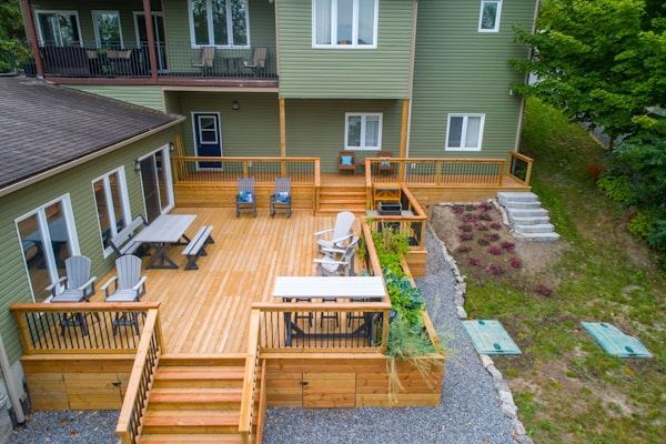 A high-angle view of a multi-level wooden deck attached to a green house, featuring outdoor furniture and staircases.