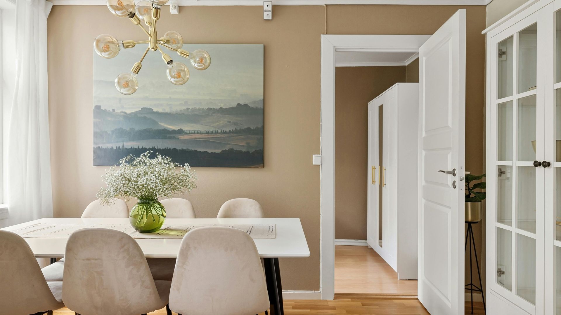 Bright dining room with white chairs, a wooden table, chandelier, wall art, and open doorway to a hallway