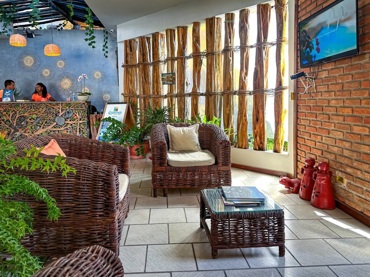 Saguão aconchegante do hotel com móveis de vime, parede com detalhes em madeira e parede de tijolos. Duas pessoas atrás de uma recepção com design exclusivo são visíveis. Pousada Pitagnda Prado