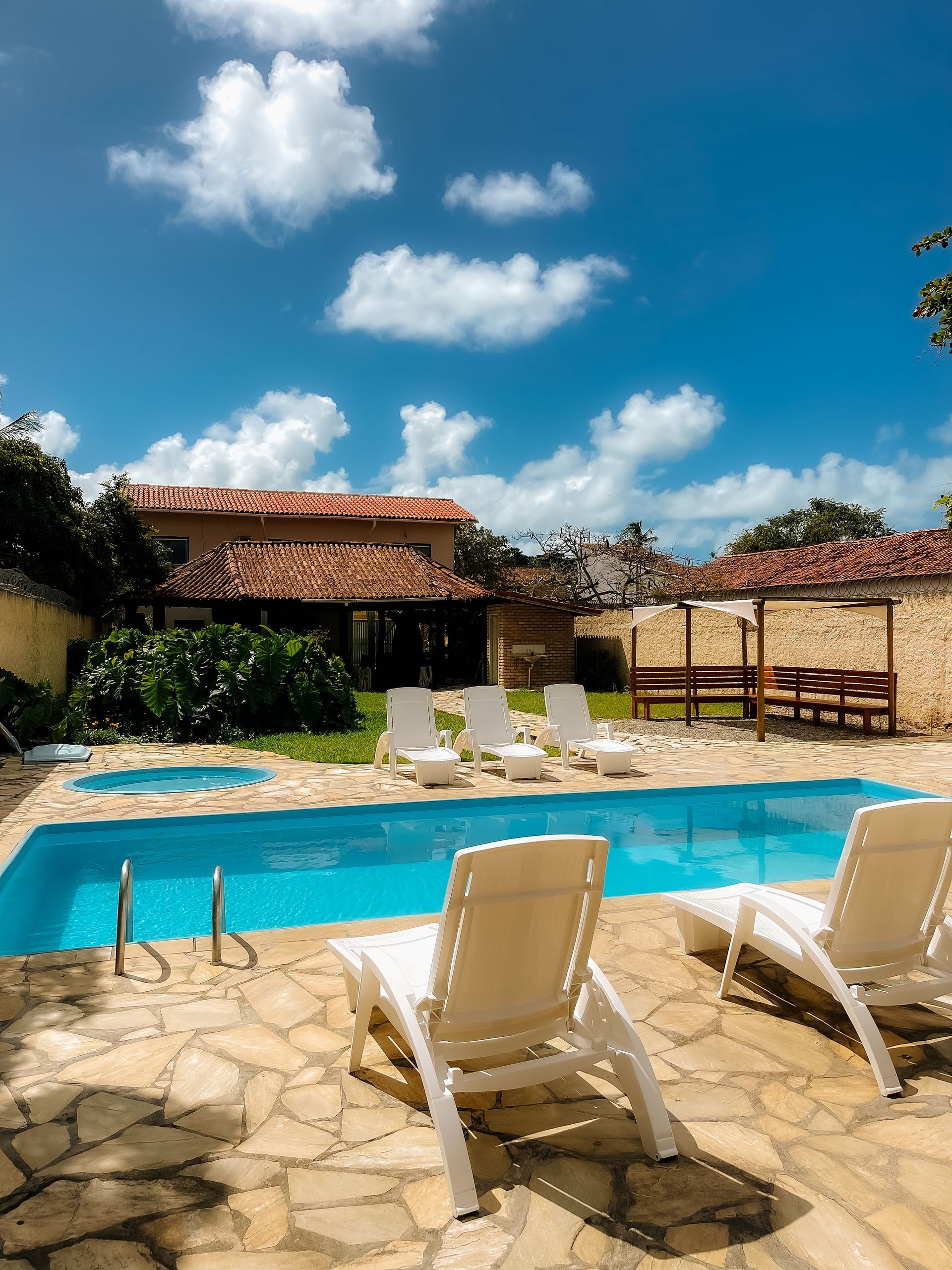 Vista da piscina de uma casa com uma piscina azul, espreguiçadeiras brancas e um céu ensolarado. Pousada Pitagnda Prado