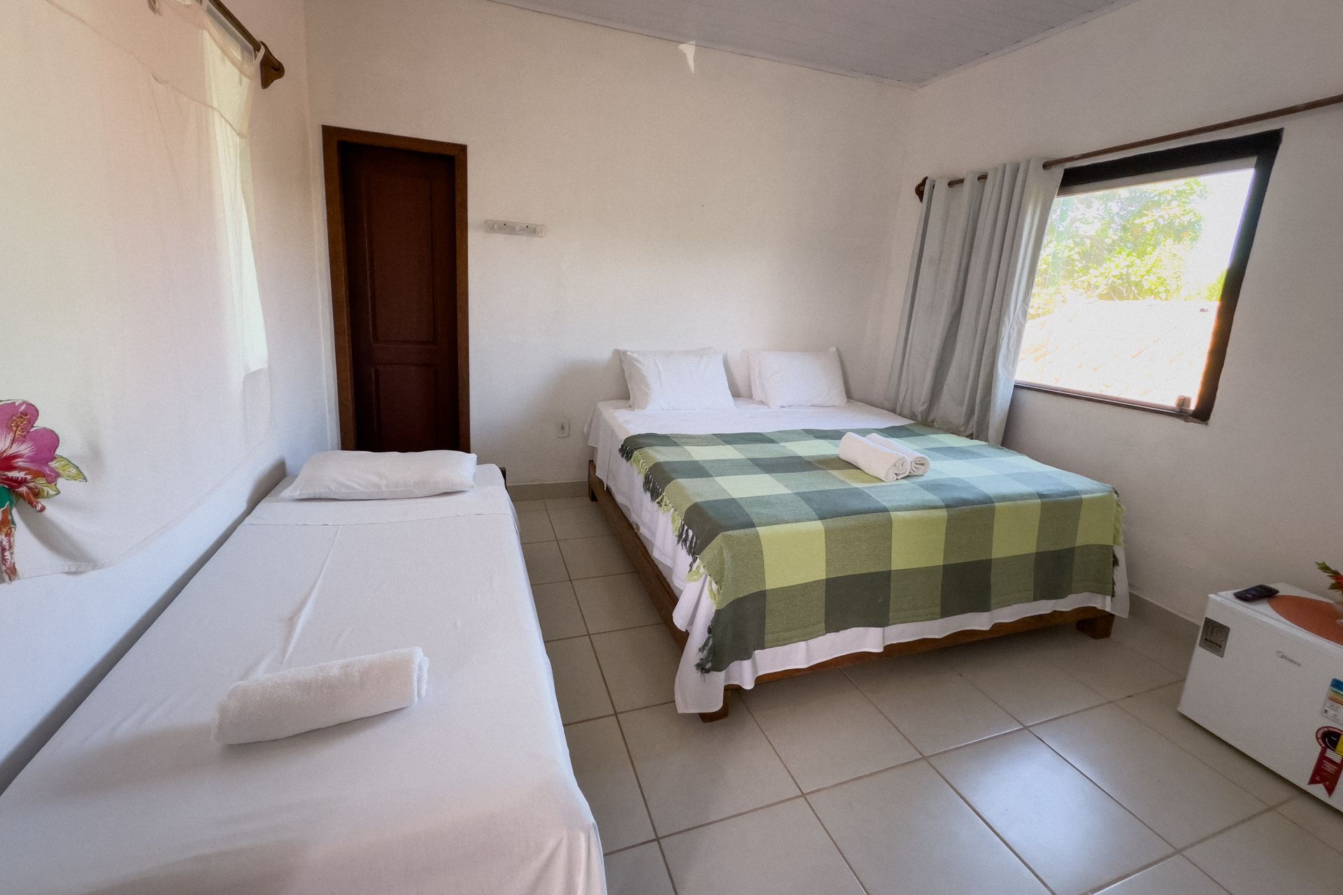 Um quarto bem iluminado com uma cama de casal e uma cama de solteiro; uma janela deixa entrar luz natural. Pousada Pitagnda Prado