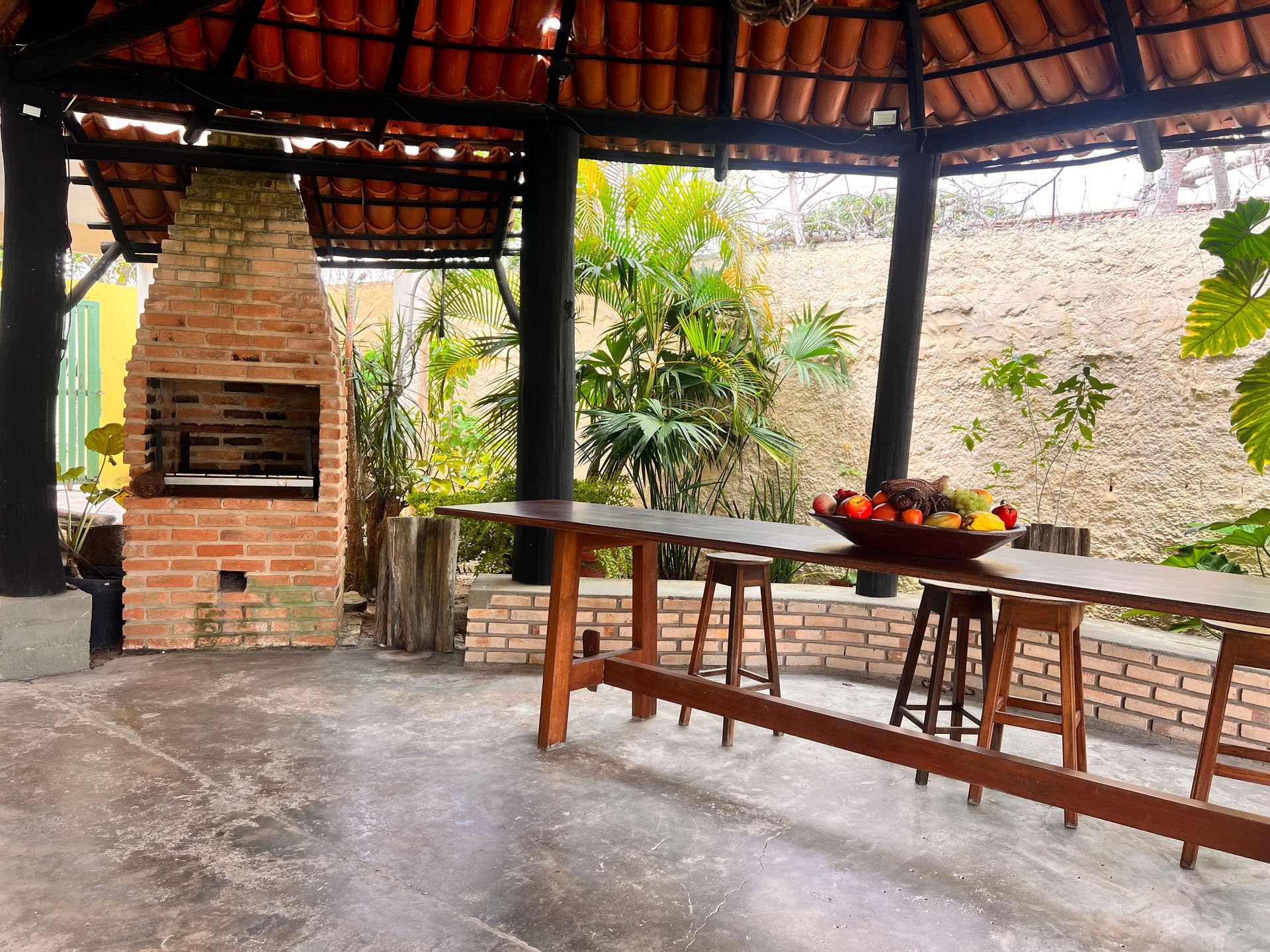 Pátio externo com churrasqueira de tijolos, mesa longa de madeira, bancos e fruteira sob um telhado de telhas.Pousada Pitagnda Prado
