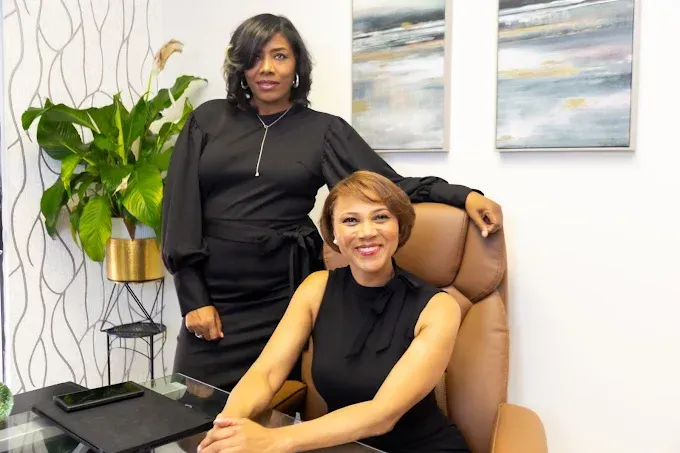 Two women in black dresses in an office setting; one seated, one standing, smiling.