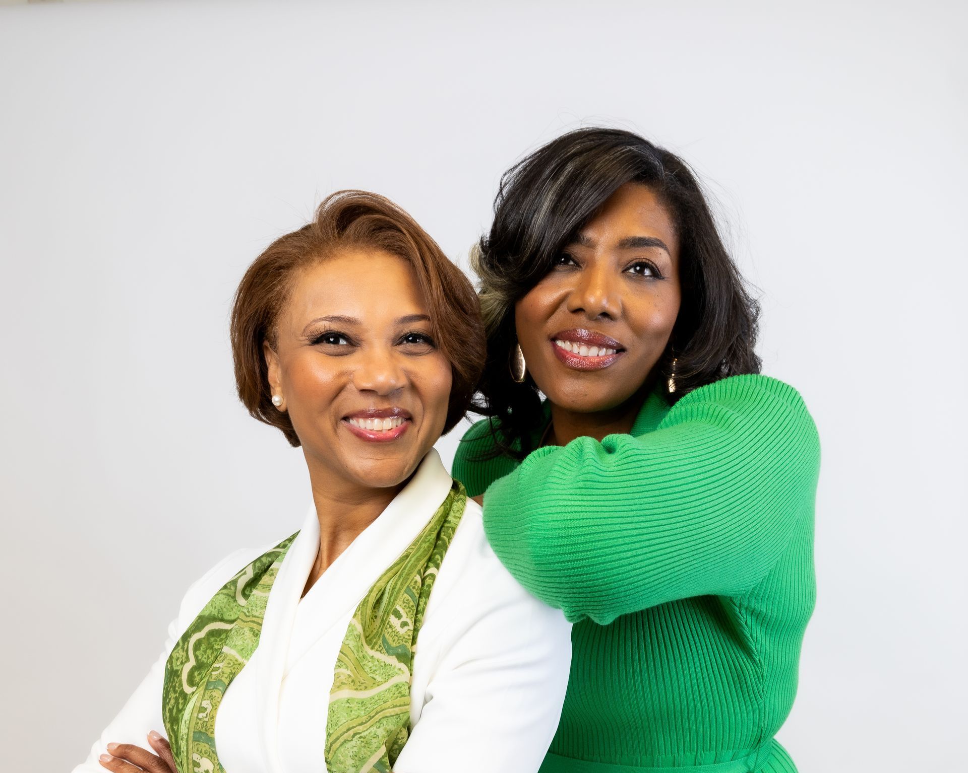 Two women, one in white blazer with green scarf, the other in green sweater, smiling and posed.