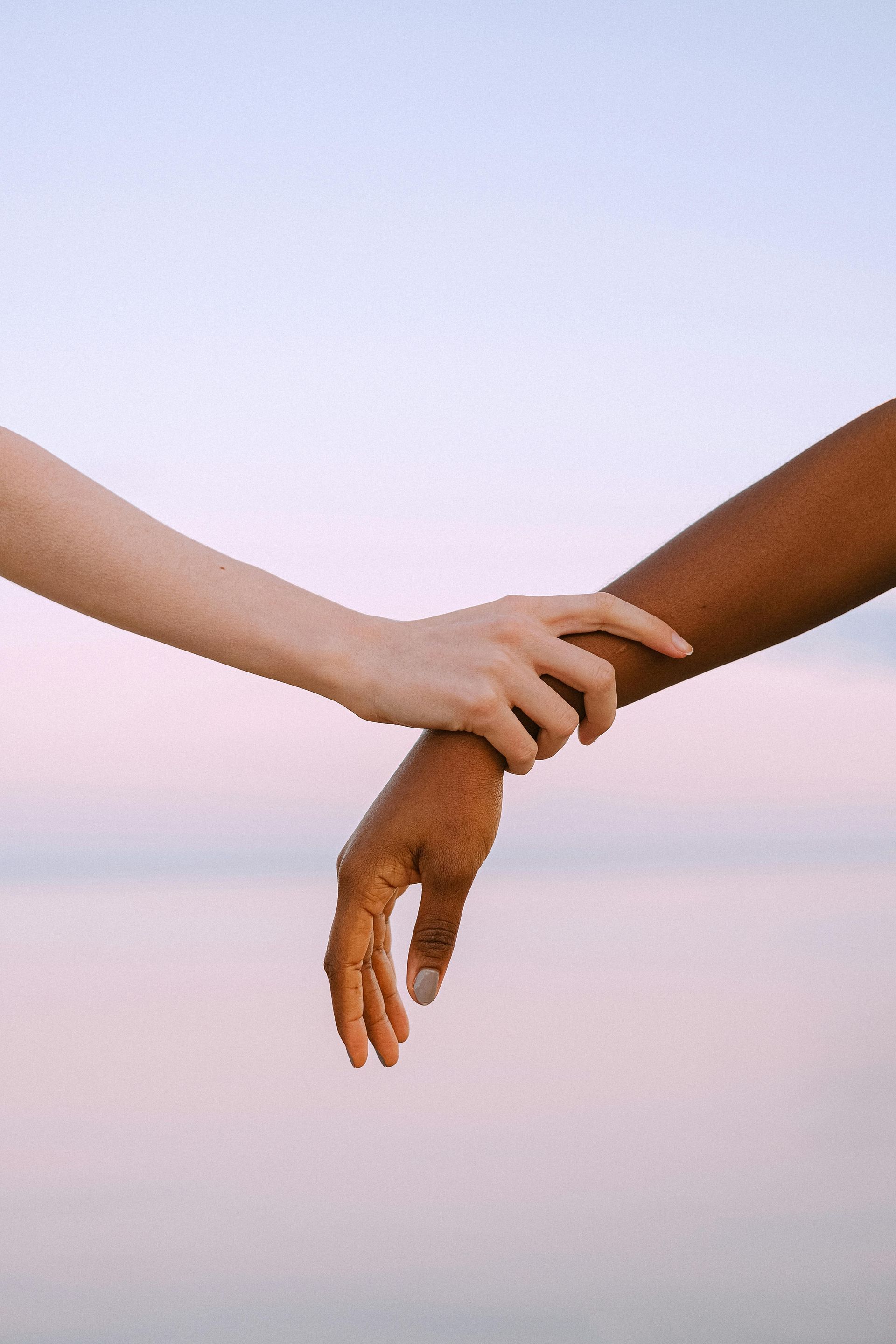 Two hands, one light-skinned and one dark-skinned, clasped in a gesture of support against a pink sky.