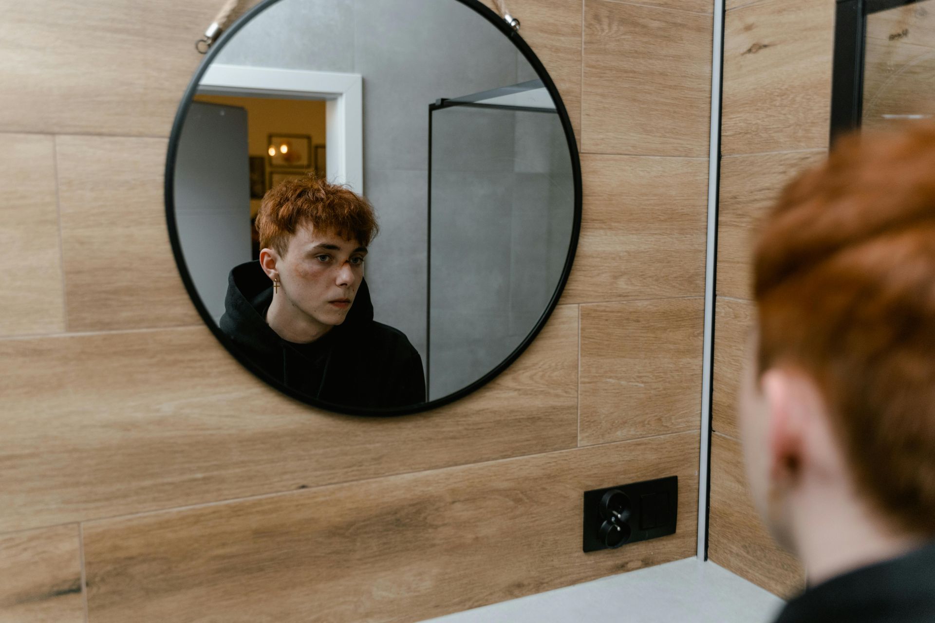 Man with red hair looking in a round mirror, wearing a black hoodie, in a bathroom.