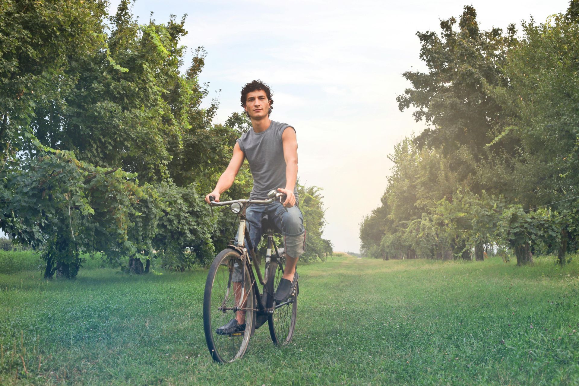 Man rides a bicycle on a grassy path lined with trees. He wears a grey tank top and shorts, looks forward.