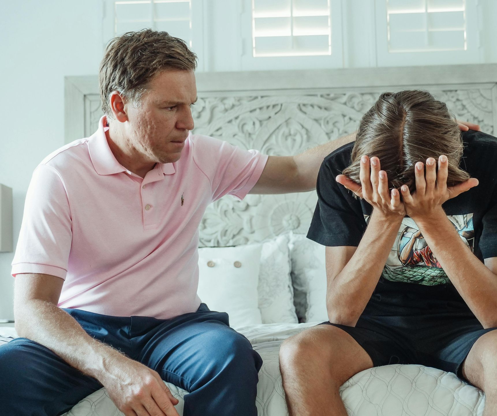 Father comforting a crying teenage son on a bed. Pink shirt, black tee, neutral bedroom.