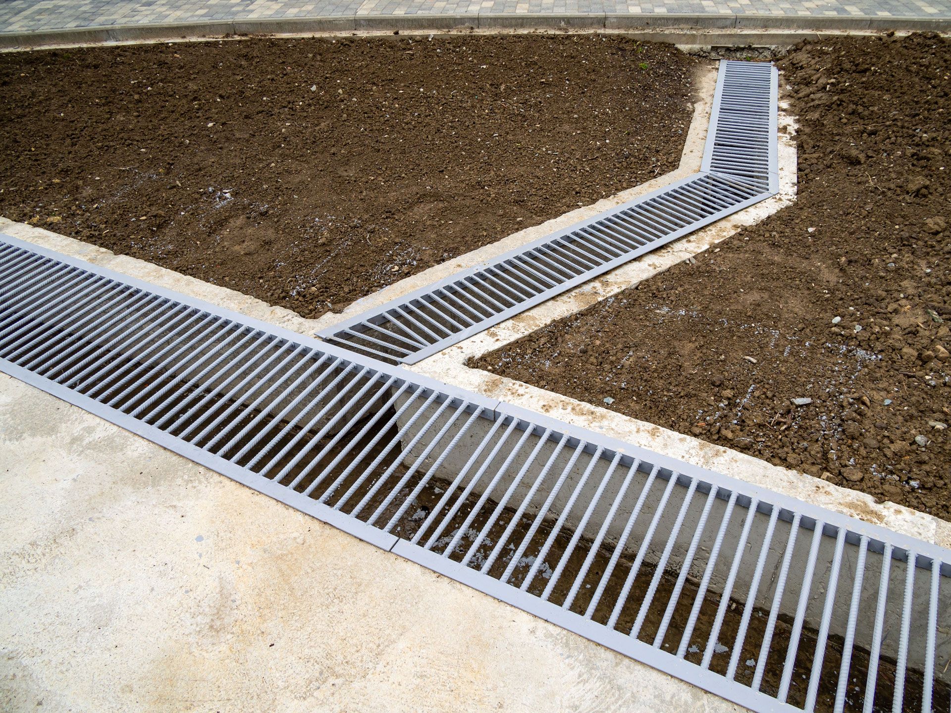 A metal trench drain grate installed in the ground between a concrete sidewalk and an area of dirt.