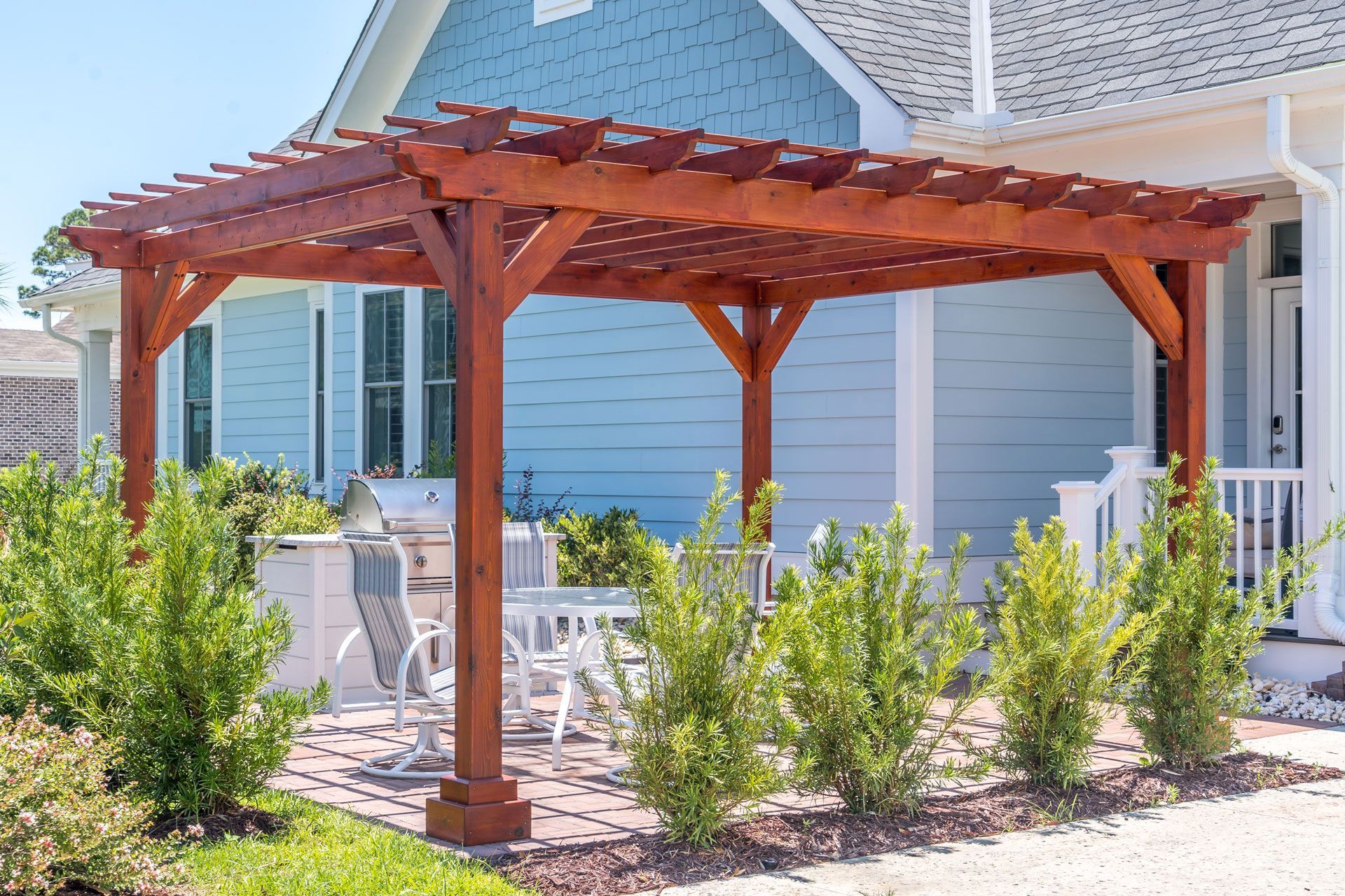 A wooden pergola stands on a brick patio beside a light blue house, shading outdoor furniture and surrounded by greenery.