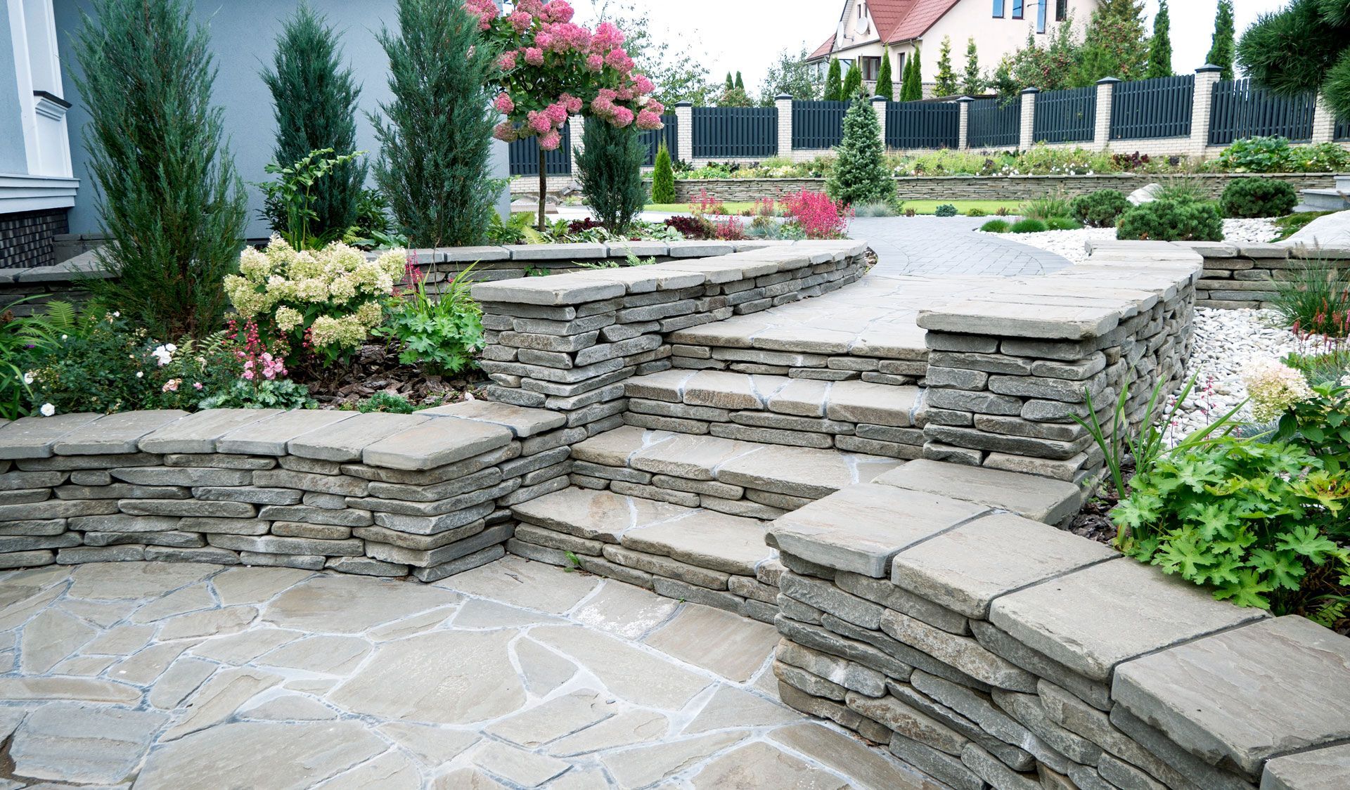 A stone patio with a curved retaining wall and steps leading to a pathway, surrounded by greenery and landscaping.