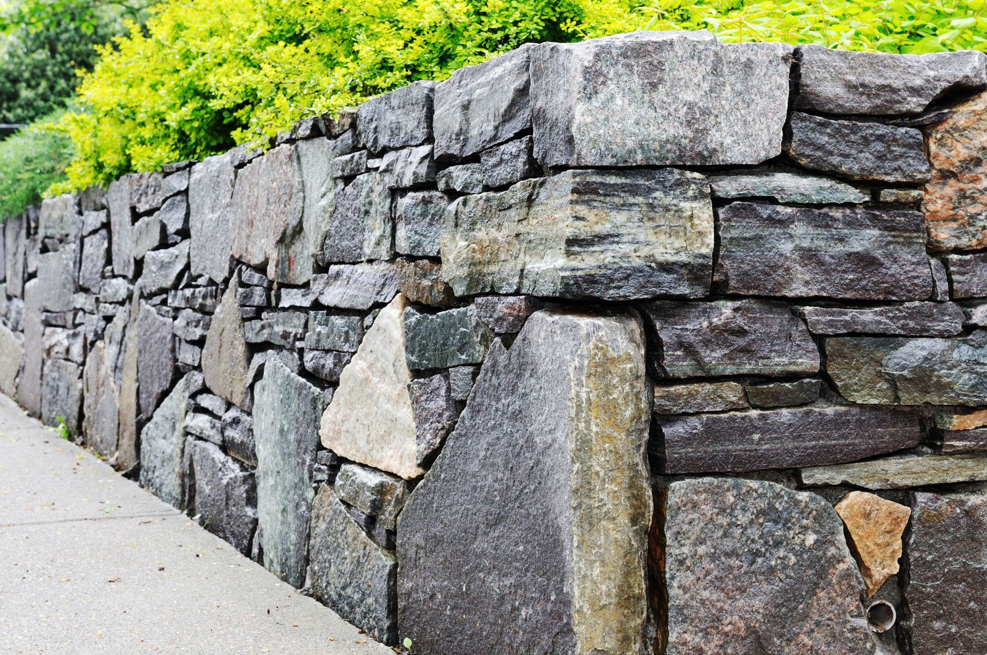 A sturdy stone wall made of varied, dark grey textured blocks borders a concrete sidewalk, with green foliage above.