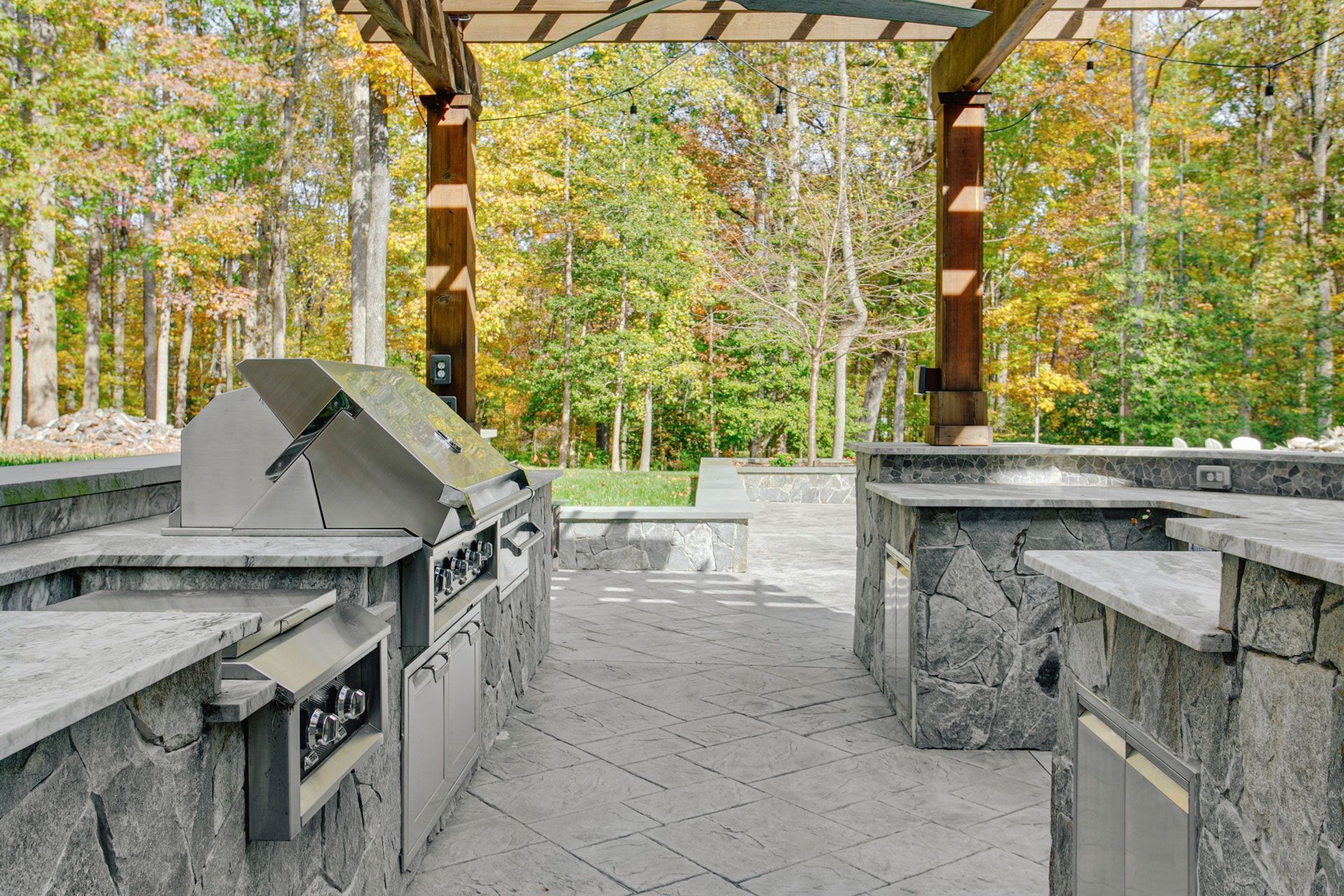 Outdoor stone kitchen with a stainless steel grill under a wooden pergola, set against a background of autumn trees.