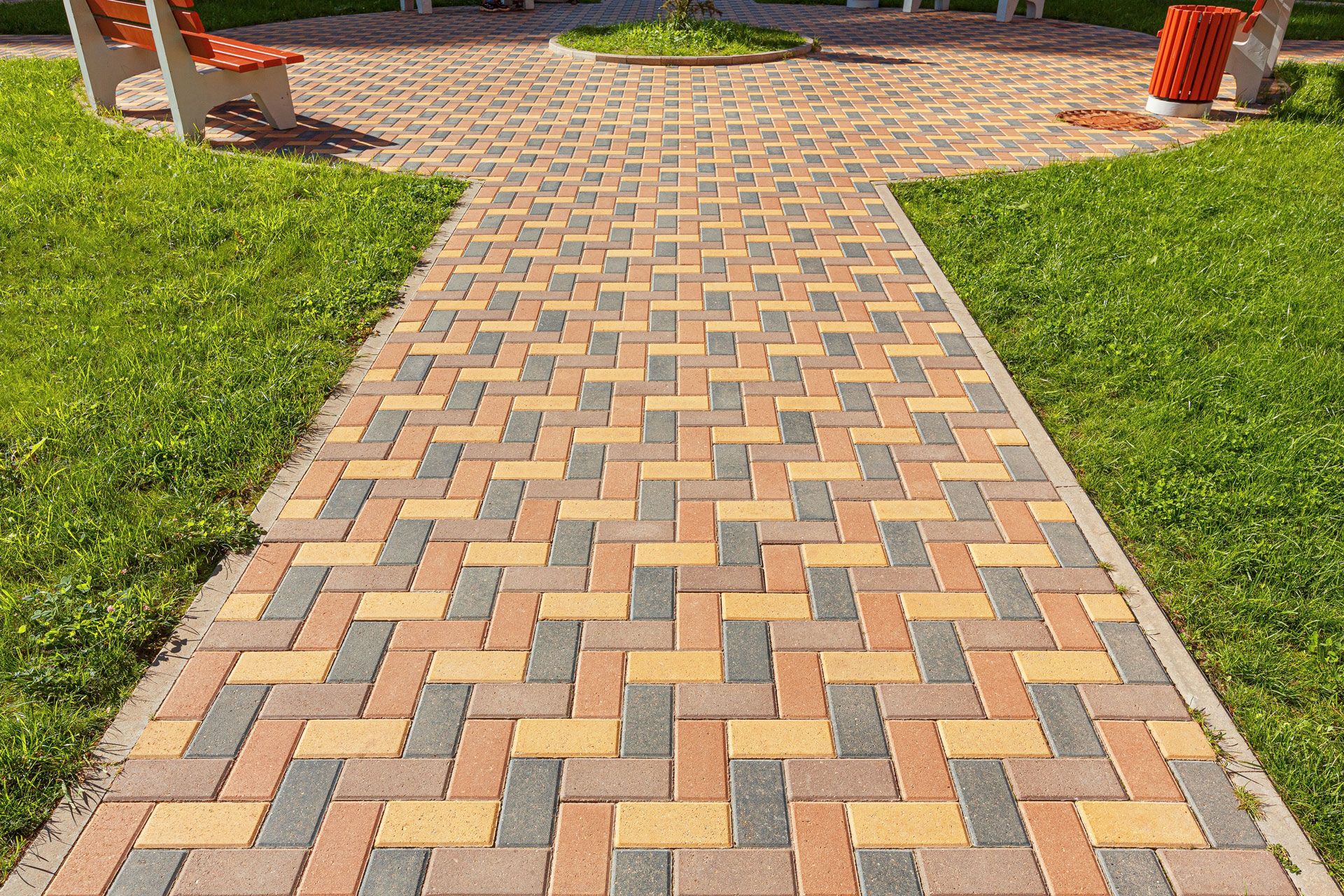 A herringbone-patterned walkway made of multicolored pavers, flanked by green grass with park benches in the background.