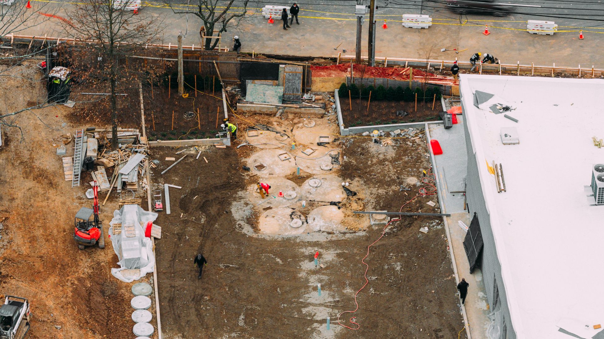 An aerial view of a construction site with excavators, exposed soil, circular concrete structures, and adjacent buildings.