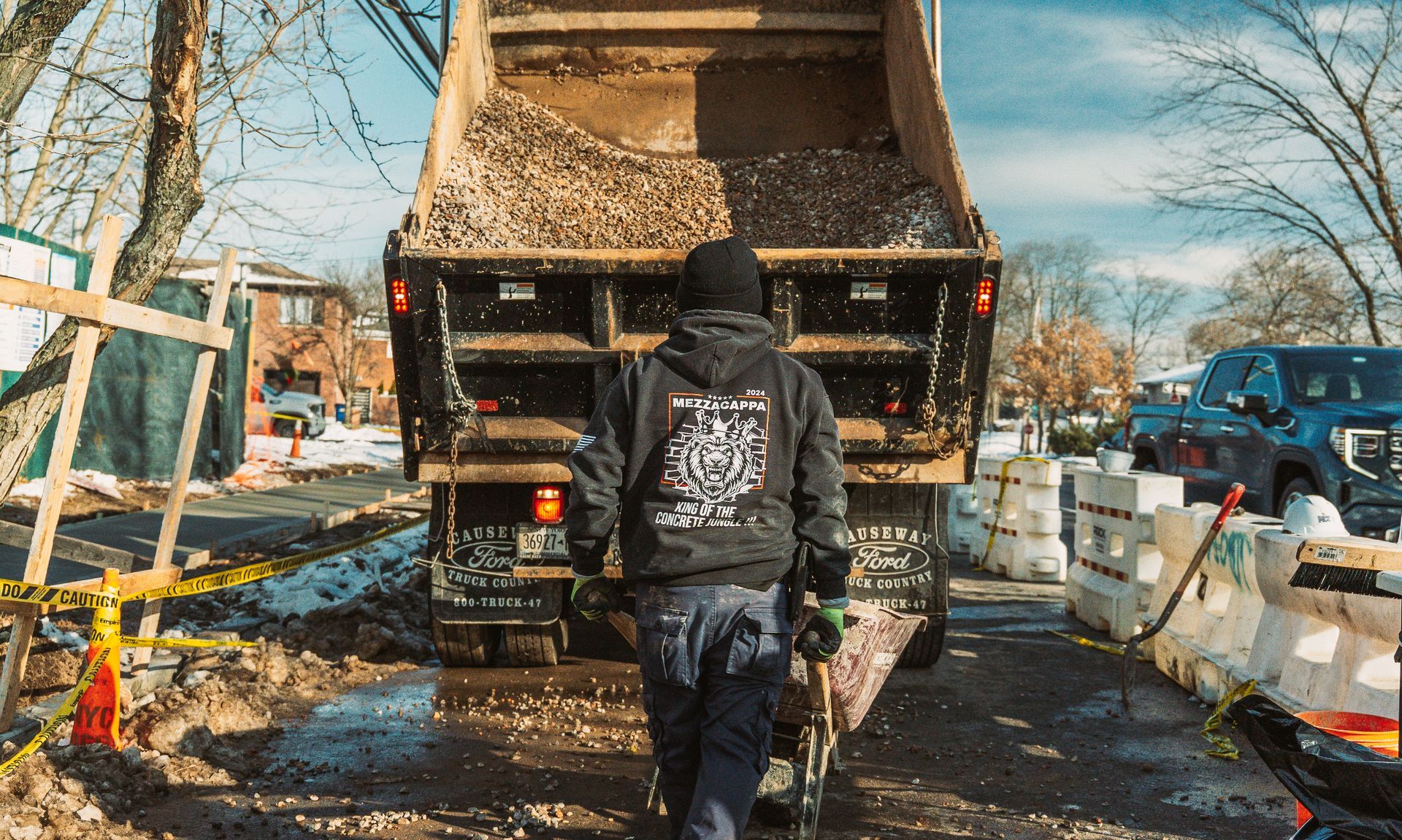 A person in a black jacket and jeans walks toward the back of a dump truck filled with debris at a construction site.