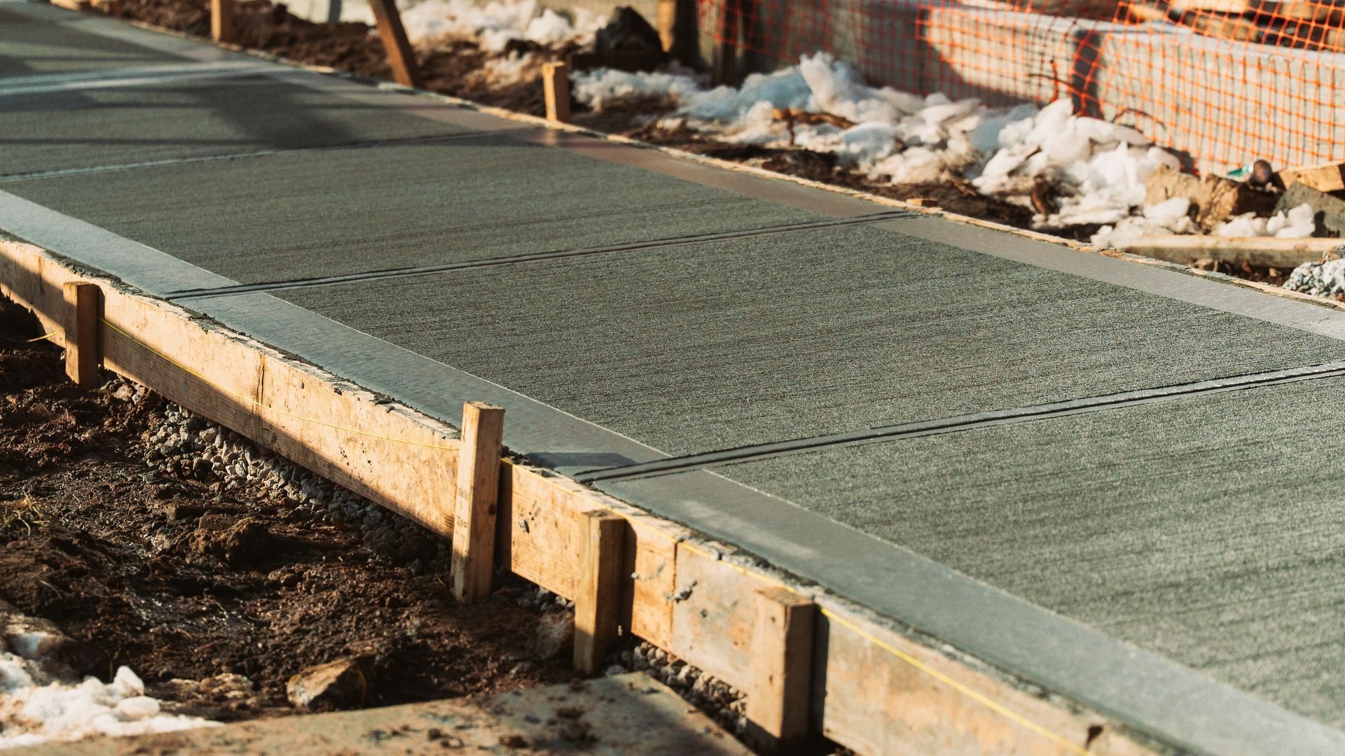 A freshly poured concrete sidewalk section framed by wooden forms, with dirt and snow patches along the edges.