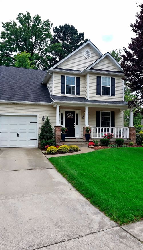 A two-story suburban house with beige siding, a dark shingled roof, a front porch, and an attached single-car garage.
