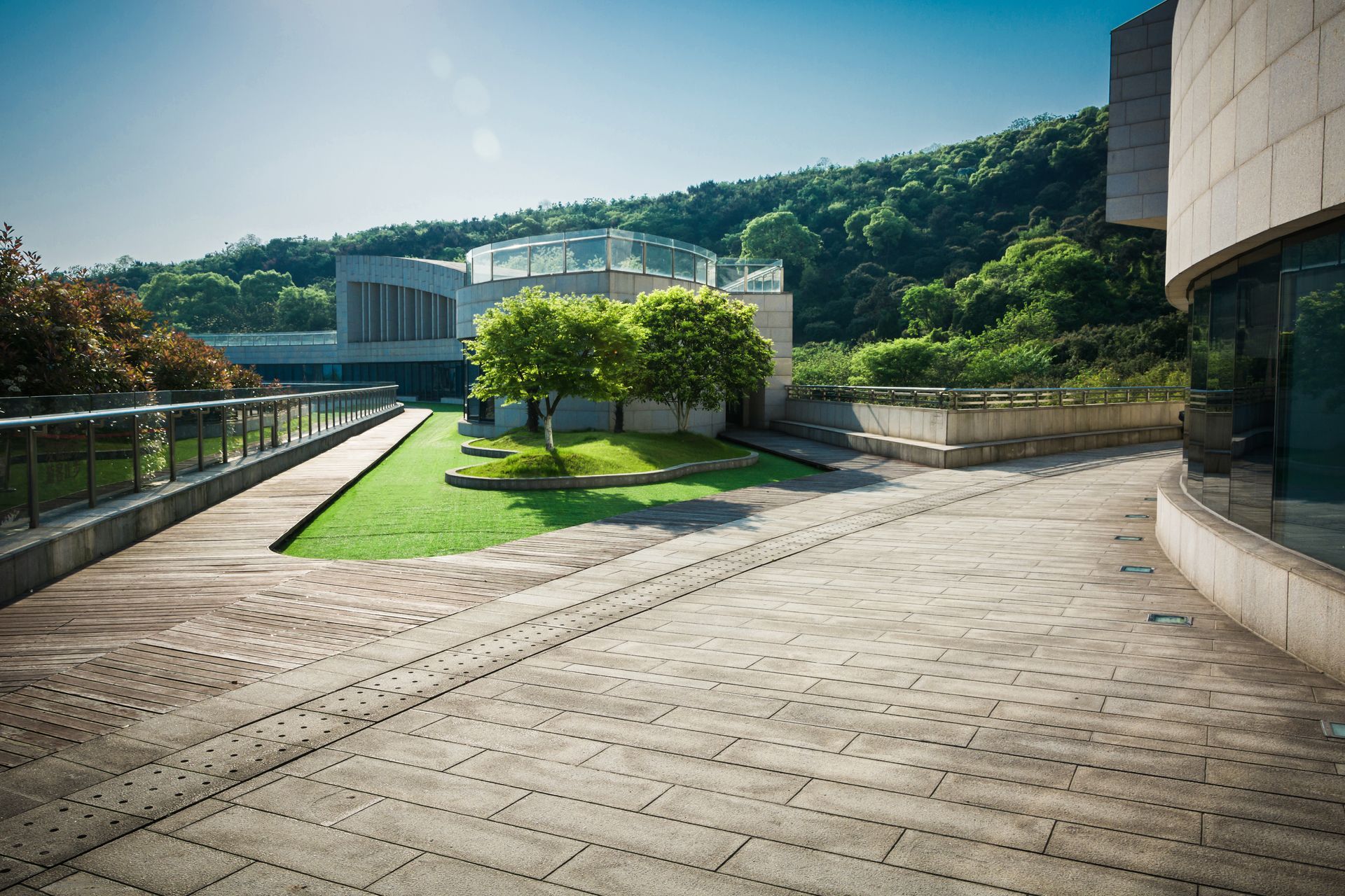 A paved outdoor courtyard leads to a small lawn with green trees, set against a backdrop of buildings and a wooded hill.