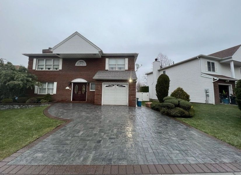 A two-story brick home with a large, patterned grey paver driveway and a white garage door under an overcast sky.