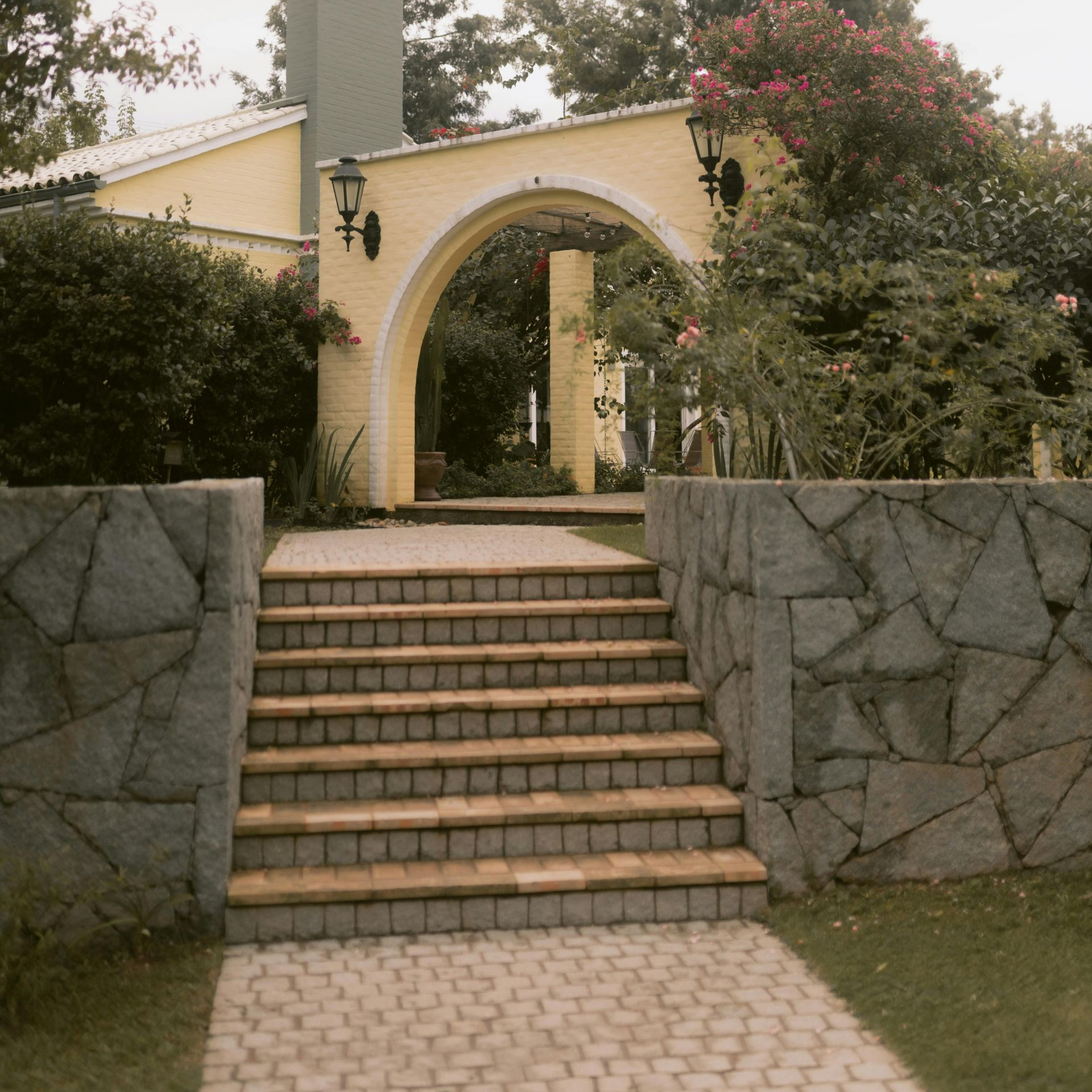 Stone stairs lead up to a yellow archway entrance of a building, framed by grey stone walls and lush green landscaping.