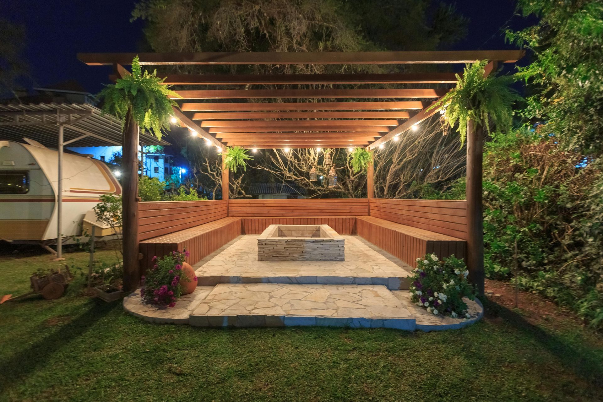 A stone fire pit area with built-in wooden benches under a pergola, decorated with hanging plants and string lights at night.