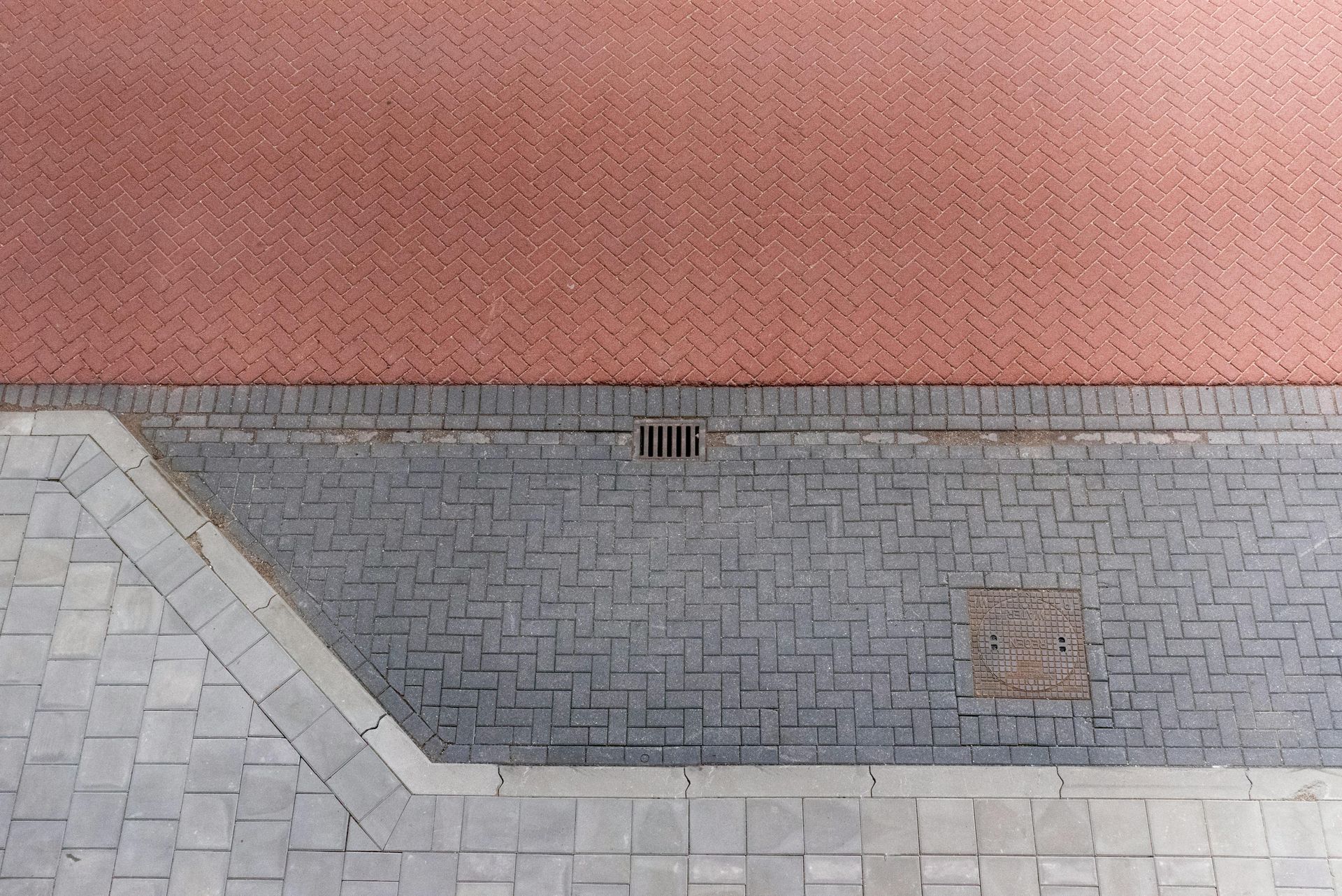 Overhead view of a sidewalk featuring red and gray paved sections, a drain grate, and a square utility cover.
