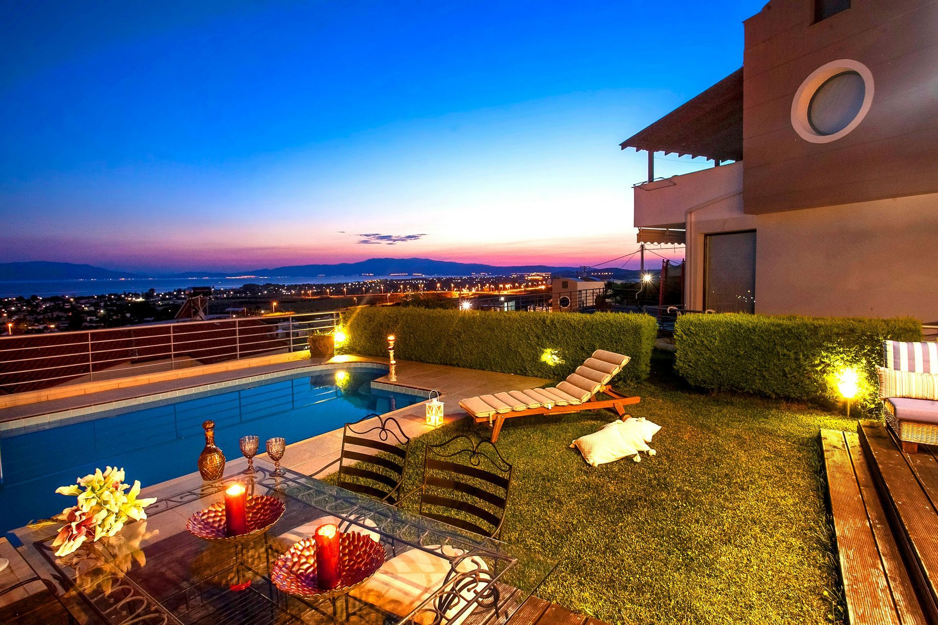 A sunset view of a house patio featuring an infinity pool, lounge chair, and dining table overlooking a city and sea.