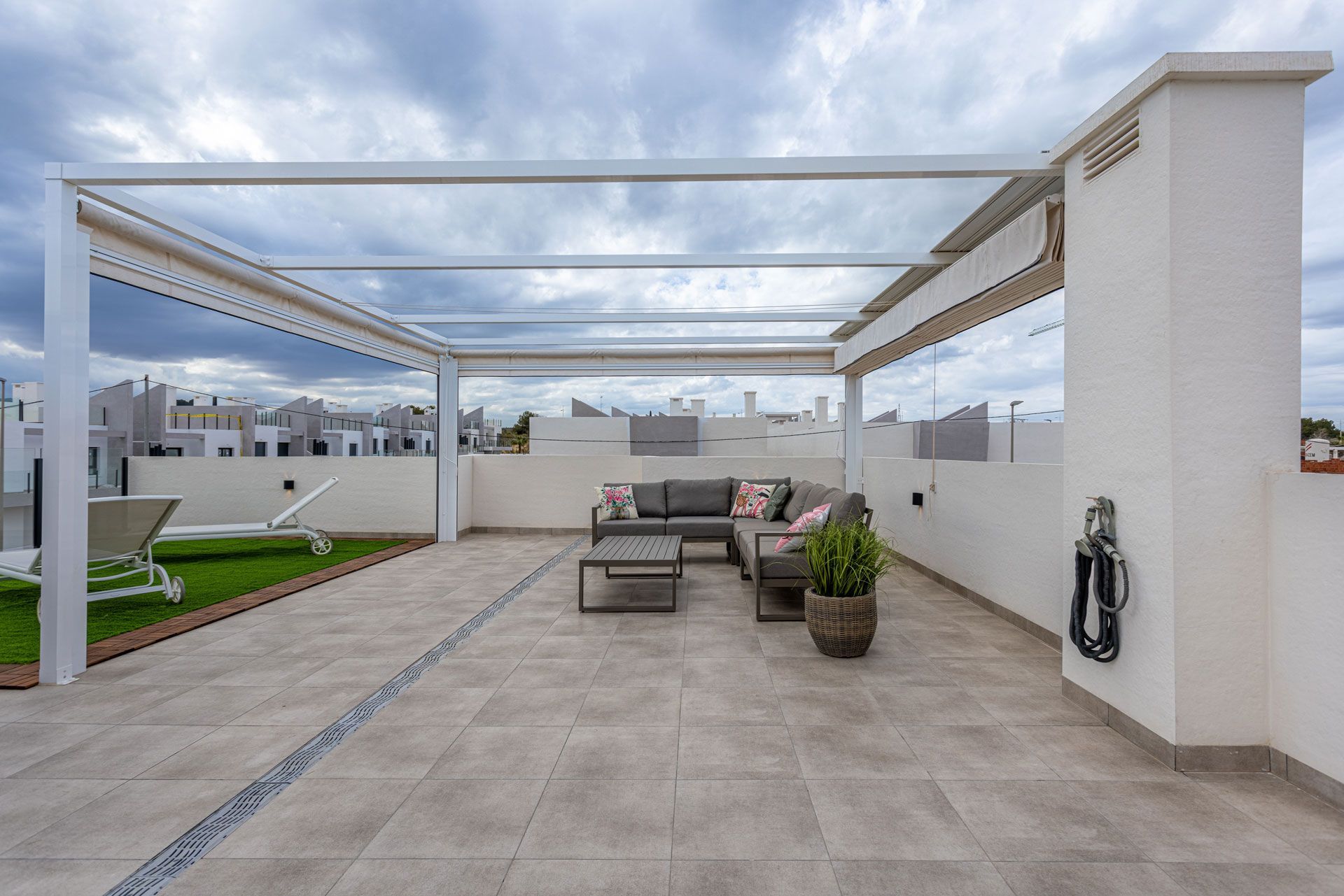 A spacious rooftop terrace featuring a modern pergola, comfortable patio furniture, potted plants, and artificial grass.