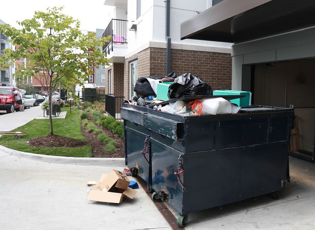Image of a black dumpster filled with rubbish bags and cardboard boxes on the driveway of a home