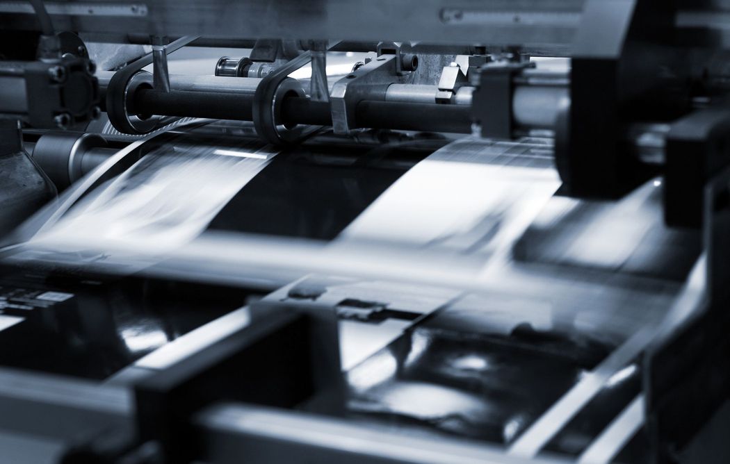 In una fabbrica, una macchina da stampa produce fogli colorati, con rotoli di carta e inchiostro ben visibili.