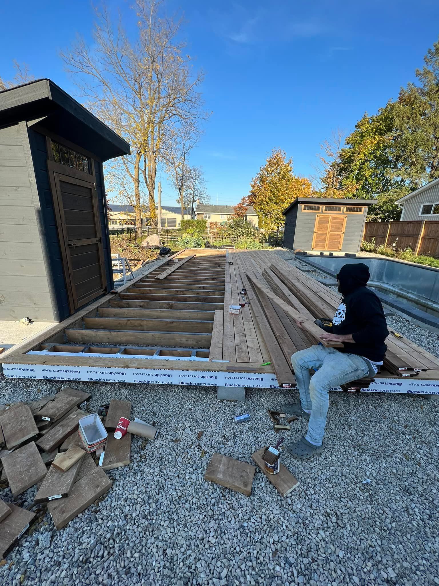 A man is sitting on a pile of wood in front of a shed.