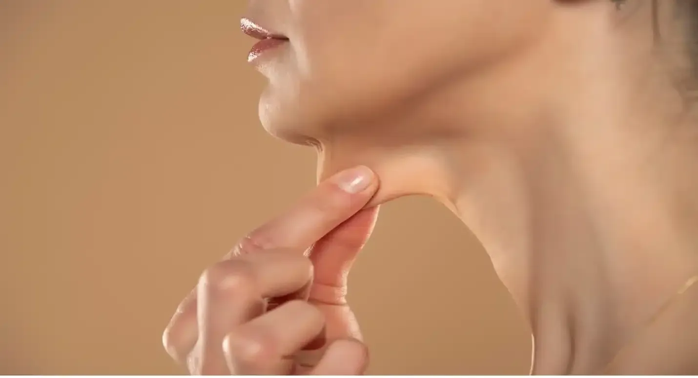 Woman pinching skin under her chin, highlighting a double chin. Beige background.